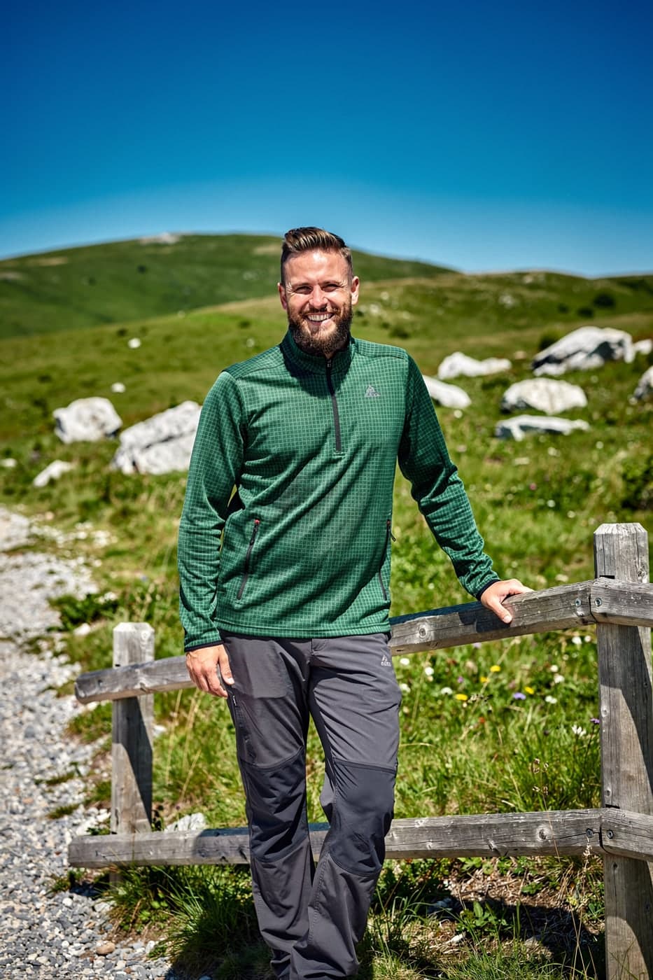 Man in a green grid fleece leaning on a wooden fence at a trailhead